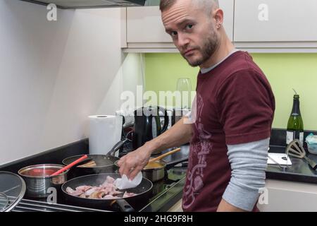 Maschio in cucina che cucina la colazione un candid shot con pentole e padelle sul piano cottura pancetta salsiccia fagioli hash brown, ammollando l'umidità in eccesso in padella Foto Stock