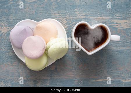 Marshmallows rotondi multicolore in un mucchio e una tazza bianca a forma di cuore con caffè caldo nero fumante su sfondo blu in legno, vista dall'alto. Deliciou Foto Stock