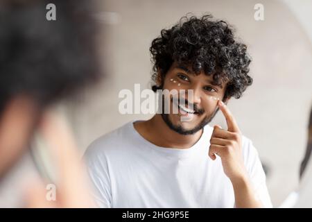 L'uomo indiano che applica la crema sotto i suoi occhi e sorridendo al suo riflesso nello specchio, godendo la routine di bellezza del mattino Foto Stock