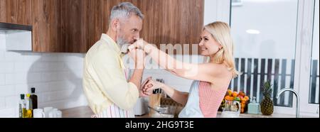 Uomo di mezza età in grembiule baciando la mano di sorridente moglie in cucina, banner Foto Stock