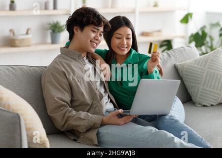 Famiglia asiatica positiva che fa shopping in linea con carta di credito e laptop, seduta sul divano e coccole a casa Foto Stock