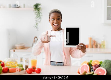 Felice emozionale giovane bella donna nera punta il dito al tablet con schermo vuoto all'interno della cucina Foto Stock