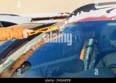 L'uomo libera l'auto arancione dalla neve. Parabrezza dell'auto. Spazzola in mano mans. Inverno, neve, veicolo. Foto Stock