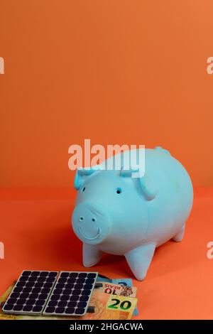 Piggy banca, pannelli fotovoltaici energia solare e denaro brasiliano isolato su sfondo arancione. Foto Stock