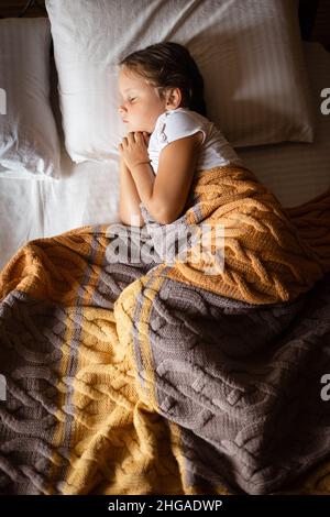 Bambina che dorme e riposa di lato con le mani piegate insieme e messo sotto il mento su un grande letto matrimoniale coperto con coperta gialla grigia indossando Foto Stock