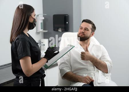 Cosmetologa femminile esperta che dà la consultazione al giovane paziente seduto su un divano in ambulatorio estetista di clinica estetica Foto Stock