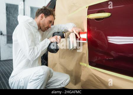 Auto Painter confronta il colore della carrozzeria dell'auto con il colore dei campioni. Uomo con una lampada di un colorista in una cabina di verniciatura di veicoli Foto Stock
