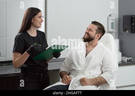 Cosmetologa femminile esperta che dà la consultazione al giovane paziente seduto su un divano in ambulatorio estetista di clinica estetica Foto Stock