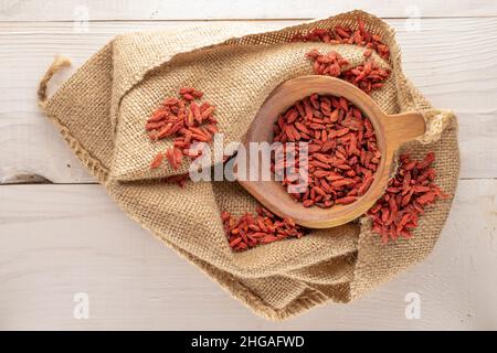 Bacche di goji biologiche essiccate in una ciotola di legno con un sacco di burlap su un tavolo di legno, macro, vista dall'alto. Foto Stock