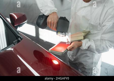 L'uomo pittore determina la tonalità corretta del colore della carrozzeria di un veicolo utilizzando una lampada speciale. Autorimessa in garage Foto Stock