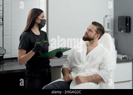 Cosmetologa femminile esperta che dà la consultazione al giovane paziente seduto su un divano in ambulatorio estetista di clinica estetica Foto Stock