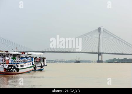 Vista sul fiume Hooghly fiancheggiato da barche ormeggiate e ponte Vidyasagar all'orizzonte sotto il cielo del mattino a Kolkata, Bengala Occidentale, India. Foto Stock