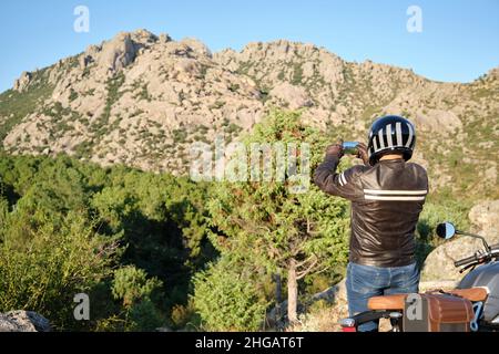 Un uomo che guida una motocicletta scattando una foto con uno smartphone in natura. Foto Stock