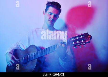 Un uomo suona una chitarra acustica in una luce neon rosso-blu. Un uomo impara a suonare la chitarra, la musica, gli hobby. Foto Stock