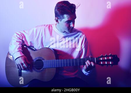 Un uomo suona una chitarra acustica in una luce neon rosso-blu. Un uomo impara a suonare la chitarra, la musica, gli hobby. Foto Stock