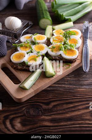 Sandwich con uova sode servito con cetriolo su un tagliere di legno con coltello Foto Stock
