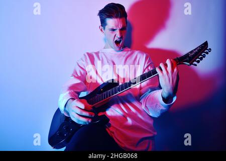 Un uomo suona la chitarra elettrica con luce al neon, luce blu rossa. Hobby, musica, club. Un uomo ama suonare la chitarra, urlare, vivide emozioni. Foto Stock