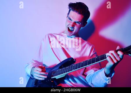 Un uomo suona la chitarra elettrica con luce al neon, luce blu rossa. Hobby, musica, club. Un uomo ama suonare la chitarra, urlare, vivide emozioni. Foto Stock