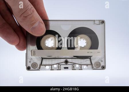 Una mano dell'uomo tiene una vecchia cassetta audio trasparente di vetro colorato per un registratore a nastro su uno sfondo bianco primo piano, la pellicola magnetica è visibile attraverso Foto Stock