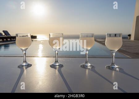 Primo piano di quattro calici con gambo di ouzo su un supporto bianco durante il tramonto con il mare Mediterraneo sullo sfondo Foto Stock