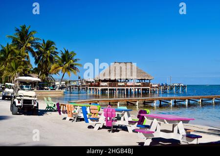 Una giornata perfetta in spiaggia con tavoli da spiaggia colorati, golf cart, moli e un palapa bar a San Pedro, Belize. Foto Stock