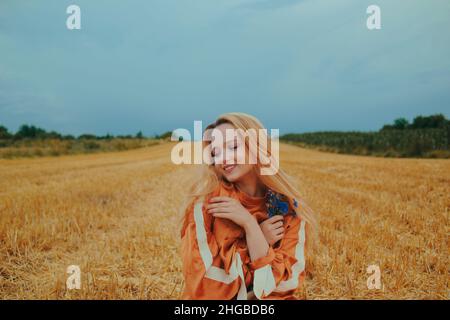 Una bella ragazza in un abito siede su un campo di grano. Ritratto fiabesco di una bionda fuori dalla città. Ragazza senza allergie tenendo fiori blu e SM Foto Stock