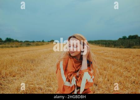 Una bella ragazza in un abito siede su un campo di grano. Ritratto fiabesco di una bionda fuori dalla città. Ragazza senza allergie tenendo fiori blu e SM Foto Stock