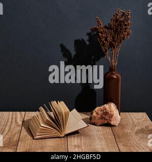 Vaso in argilla con fiori secchi marroni, pietra naturale, vecchio libro aperto su tavola di legno Foto Stock