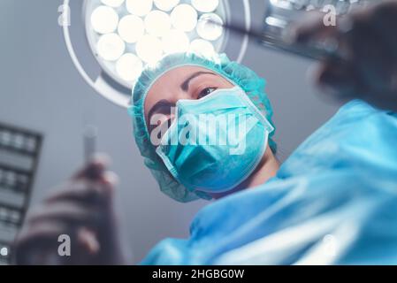 Da sotto chirurgo donna in uniforme medica con l'uso di strumenti professionali mentre si trova in piedi sotto la luce luminosa in sala operatoria. Foto di alta qualità Foto Stock