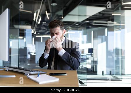 Un uomo con un naso che cola e la febbre lavora in ufficio, un uomo d'affari malato al computer ha un naso che cola Foto Stock