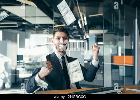 Bel giovane uomo d'affari gioioso seduto su una scrivania, lavorando in un ufficio moderno, gettando soldi sulla testa. L'uomo sorridente felice getta soldi nella A. Foto Stock