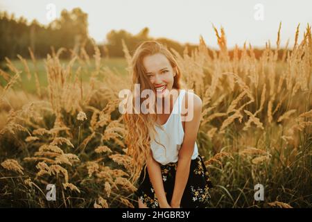 Splendida donna in un campo di grano su sfondo tramonto. Una ragazza alla moda con capelli lunghi gioisce, ride, gode di vita ed estate, natura, felicità. Foto Stock