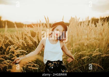 Splendida donna in un campo di grano su sfondo tramonto. Una ragazza alla moda con capelli lunghi gioisce, ride, gode di vita ed estate, natura, felicità. Foto Stock