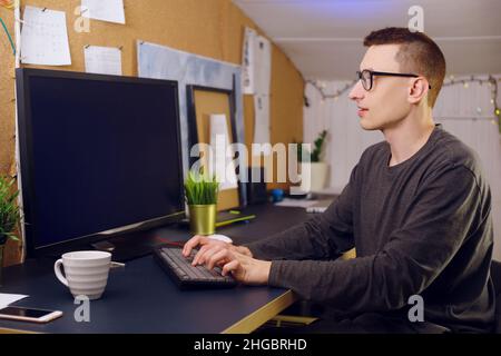 L'uomo in occhiali utilizza la tastiera e il mouse guarda lo schermo verde. Un uomo lavora a casa. Rimani a casa, freelance, videogamer. Foto Stock