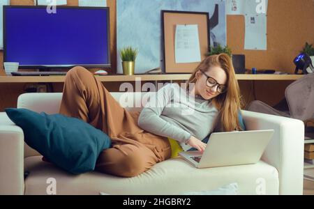 Ragazza sdraiata su un divano guardando un computer portatile. La ragazza si trova sul devan e guarda programmi TV, video, procrastinazione. Foto Stock