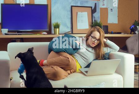 Ragazza sdraiata su un divano guardando un computer portatile. La ragazza si trova sul devan e guarda programmi TV, video, procrastinazione. Foto Stock