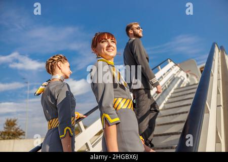 Due donne stewardesses e uomo aereo pilota salire piano di scale sotto il cielo blu Foto Stock