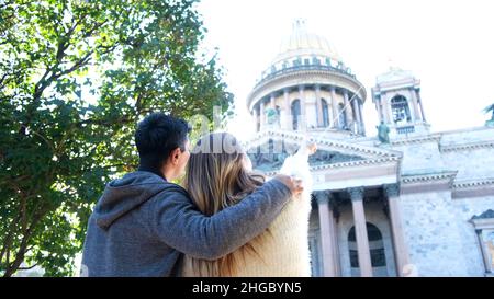 Vista posteriore di una coppia felice in piedi e discutere incredibile tempio vicino verde cespuglio. Uomo che abbraccia la donna, guardando la bella Cattedrale e indicando un f Foto Stock