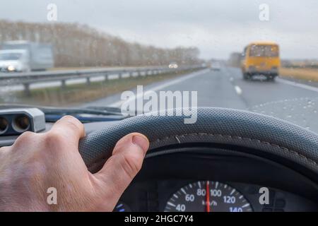 la mano del conducente si trova sul volante all'interno della vettura, che si sta guidando lungo un'autostrada suburbana, bagnata dalla pioggia Foto Stock