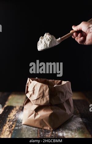 Farina di grano in un pacchetto di mestiere. Movimento di congelamento, cucchiaio sfondo scuro, spolverare la farina. Vista frontale. Foto Stock