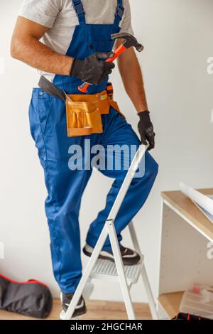 Scatto corto di elettricista in uniforme usando la scala, tenendo il martello mentre installa il raccordo leggero nel nuovo appartamento Foto Stock