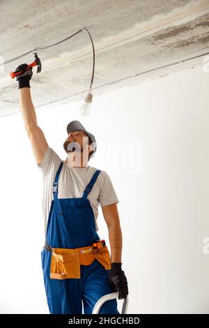 Elettricista concentrato in uniforme usando il martello mentre si collega il filo al soffitto, installando il raccordo della luce in appartamento Foto Stock