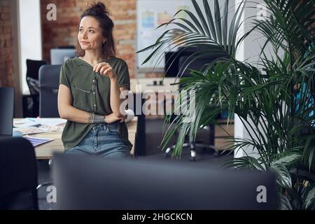 Donna di design con interni premurosi che tiene gli occhiali e guarda da parte mentre si trova in un moderno ufficio verde durante il giorno Foto Stock