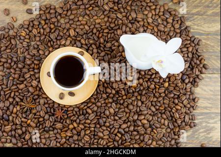 vista dall'alto di una piccola tazza di caffè nero e di una caraffa di latte in piedi sui chicchi di caffè. sfondo in legno, fiore bianco di orchidea Foto Stock