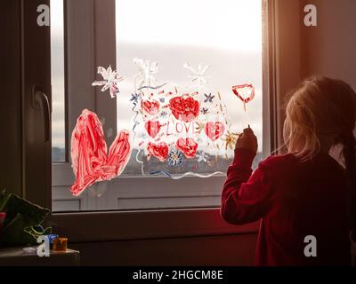 Bambina con pennello in mano disegno rosso cuore Amore sul vetro della finestra. San Valentino, sogni, speranza, quarantena famiglia tempo libero. Soggiorno a casa arte co Foto Stock