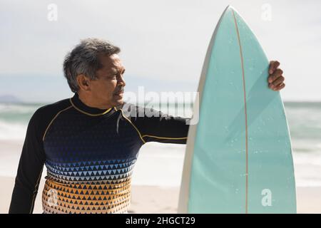 Uomo anziano biraciale che guarda via mentre si alza in piedi con la tavola da surf in spiaggia nelle giornate di sole Foto Stock