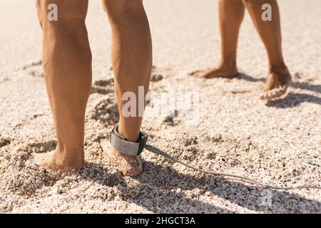 Sezione bassa dell'anziano biraciale con guinzaglio da surf sulla caviglia da donna sulla sabbia alla spiaggia soleggiata Foto Stock