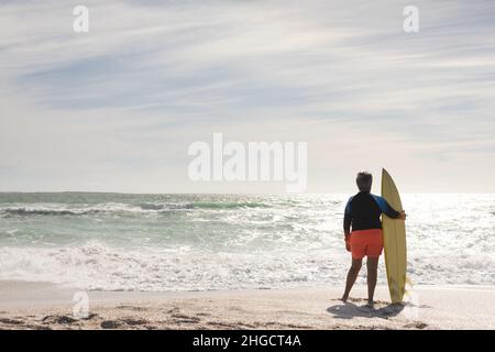 Vista posteriore a tutta lunghezza della donna biraciale anziana che tiene la tavola da surf guardando l'orizzonte dalla spiaggia soleggiata Foto Stock