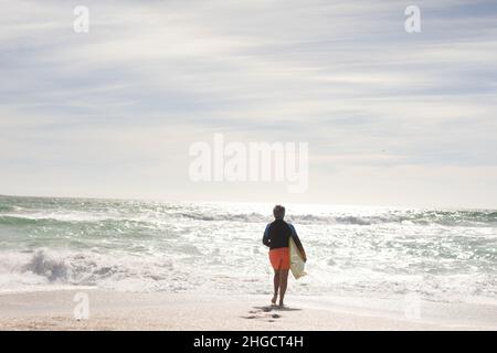 Vista posteriore della donna biraciale anziana che tiene la tavola da surf che corre verso il mare in spiaggia il giorno di sole Foto Stock