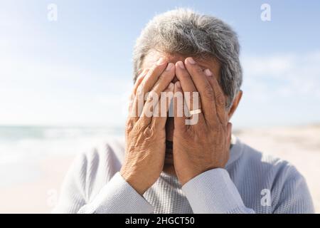 Primo piano di un uomo anziano biraciale preoccupato che copre il viso con le mani in spiaggia contro il cielo nelle giornate di sole Foto Stock